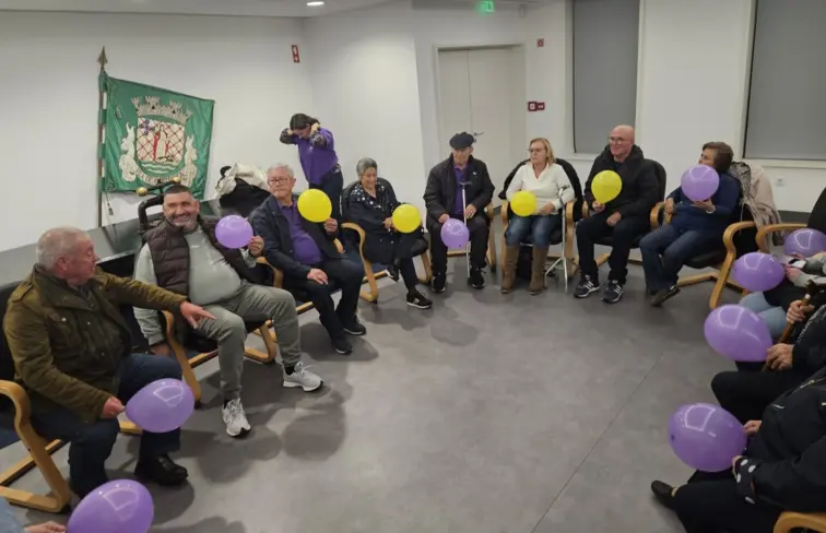 Doze pessoas, de várias idades, sentadas em cadeiras, em meia-lua, metade está de costas e as restantes de perfil, numa sala branca, com uma televisão onde está escrito "Obrigado", ao lado está uma mulher de meia-idade de pé, em frente ao grupo e com um 